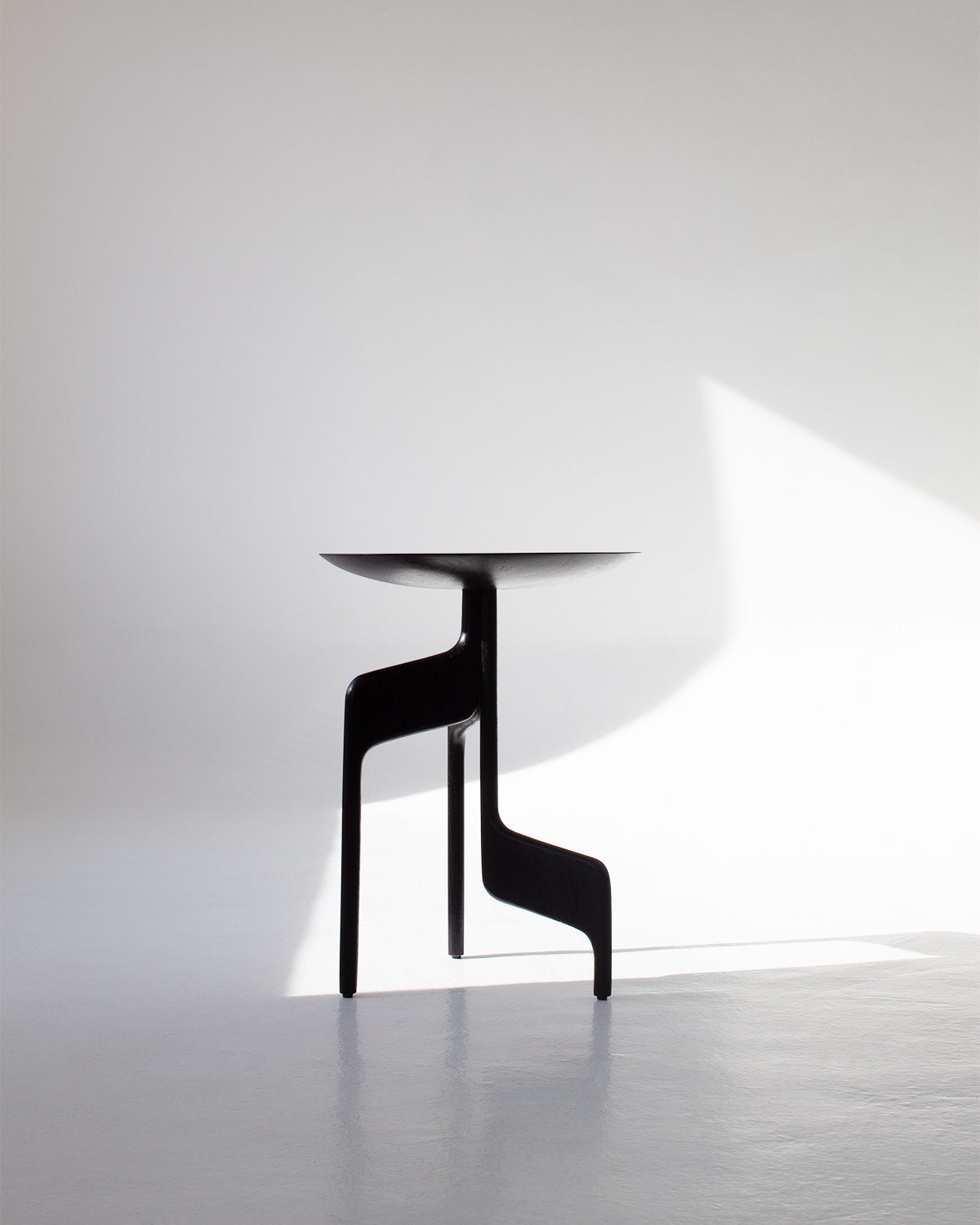 The "Vereda" Side Table by Luciano Costa Estúdio, featuring modern black angled legs, stands in a bright minimalist room as sunlight casts geometric shadows on the white floor and wall.
