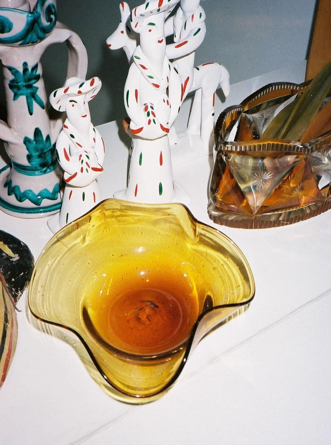 The Michele Bowl Small by Akua Objects, a wavy yellow Venetian glass piece, sits on a white surface near ceramic figurines in dotted white clothing and a faceted glass box with gold utensils inside.