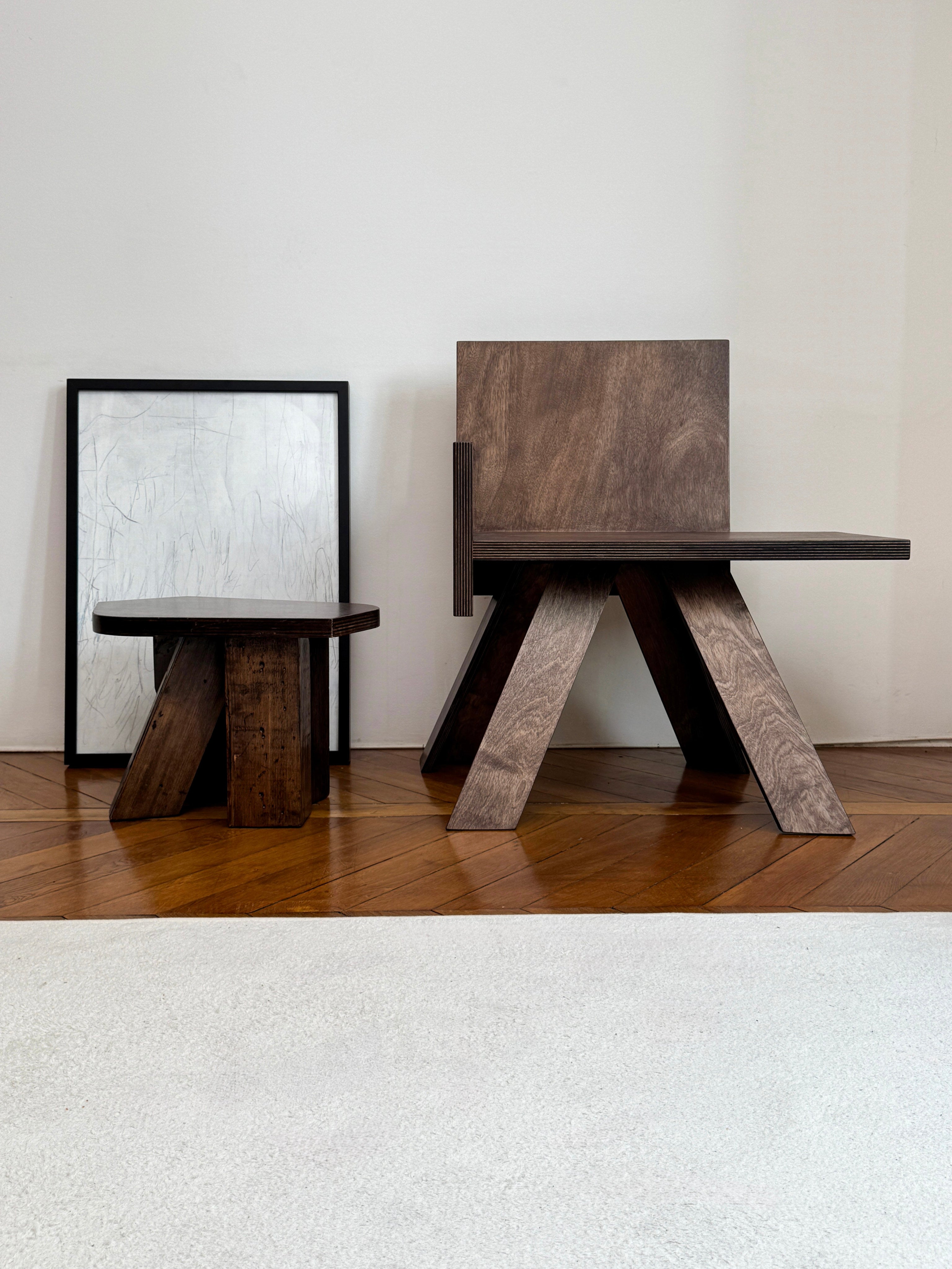 Two GOONS End Table Chairs with geometric legs sit on a wooden floor next to framed abstract art and a light rug, showcasing the brand’s artisanal design against a white wall.