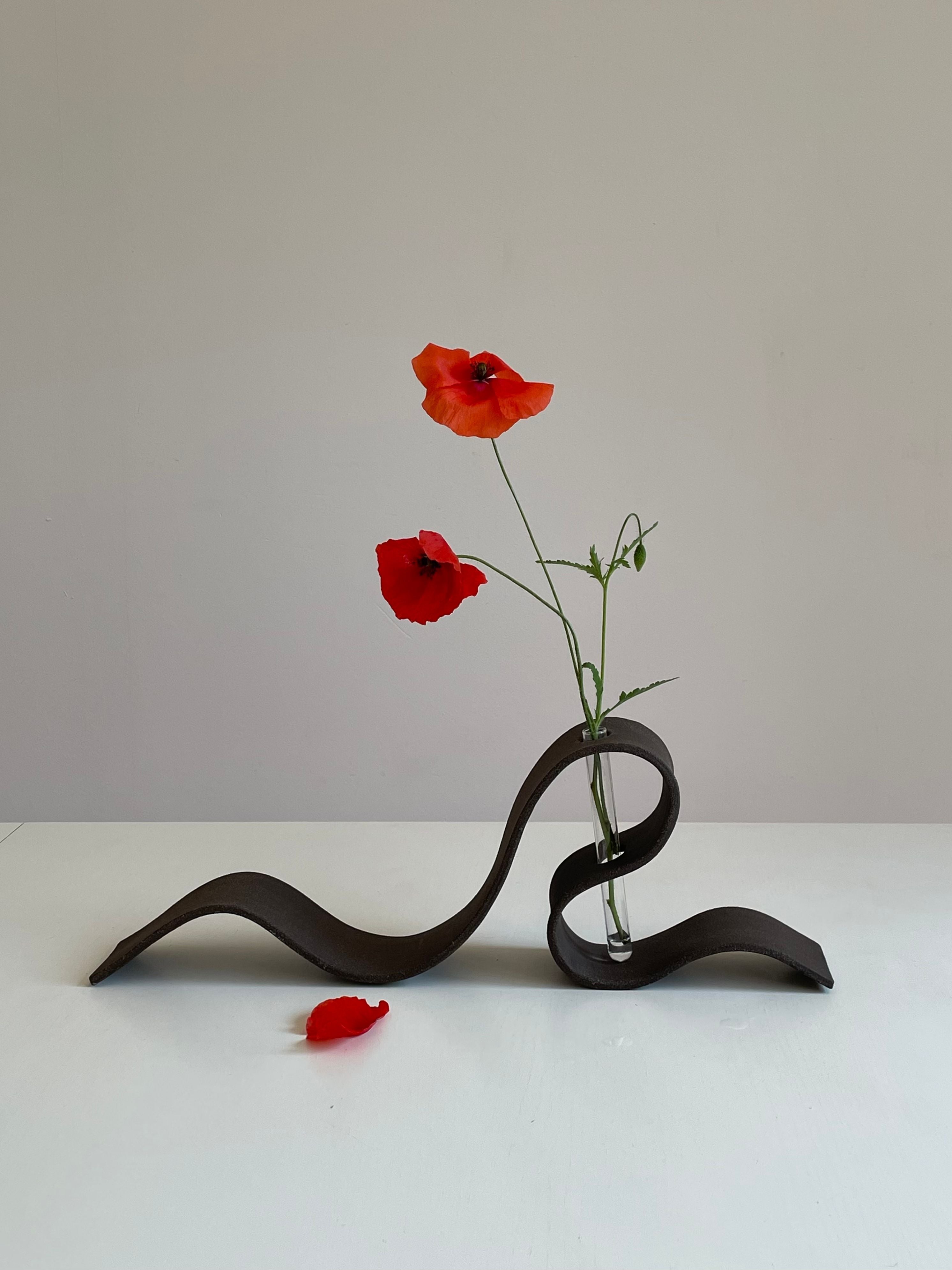 The LESS Ribbon Vase in Black features a minimalist wavy design holding a glass tube with two red poppies, one petal fallen on the white surface below against a plain light background.