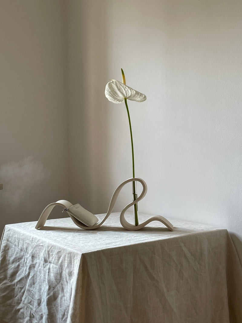 A single white anthurium in the LESS Ribbon Vase in Off White sits on a textured, light cloth-covered table against a softly lit, plain wall.