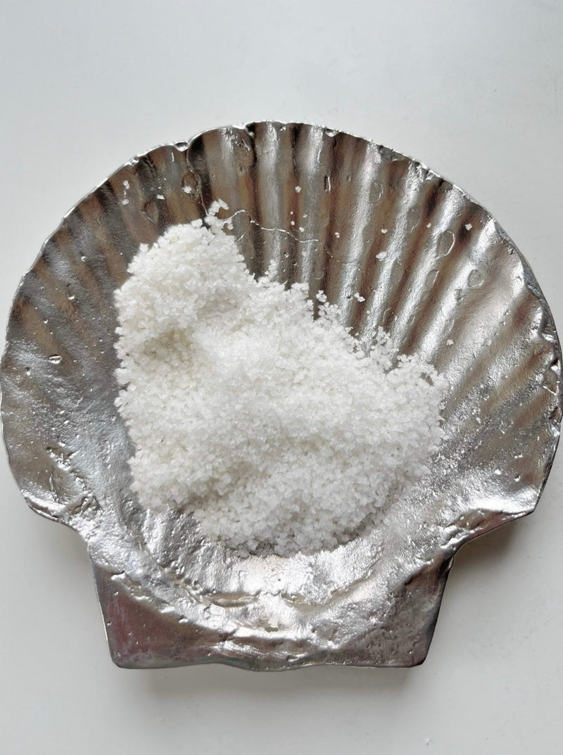 A mound of coarse white salt sits atop Médecine's Fossil Shell Plate in Silver, its hand-forged, scallop-shaped design elegantly highlighting the beauty of decorative tableware against a clean white background.