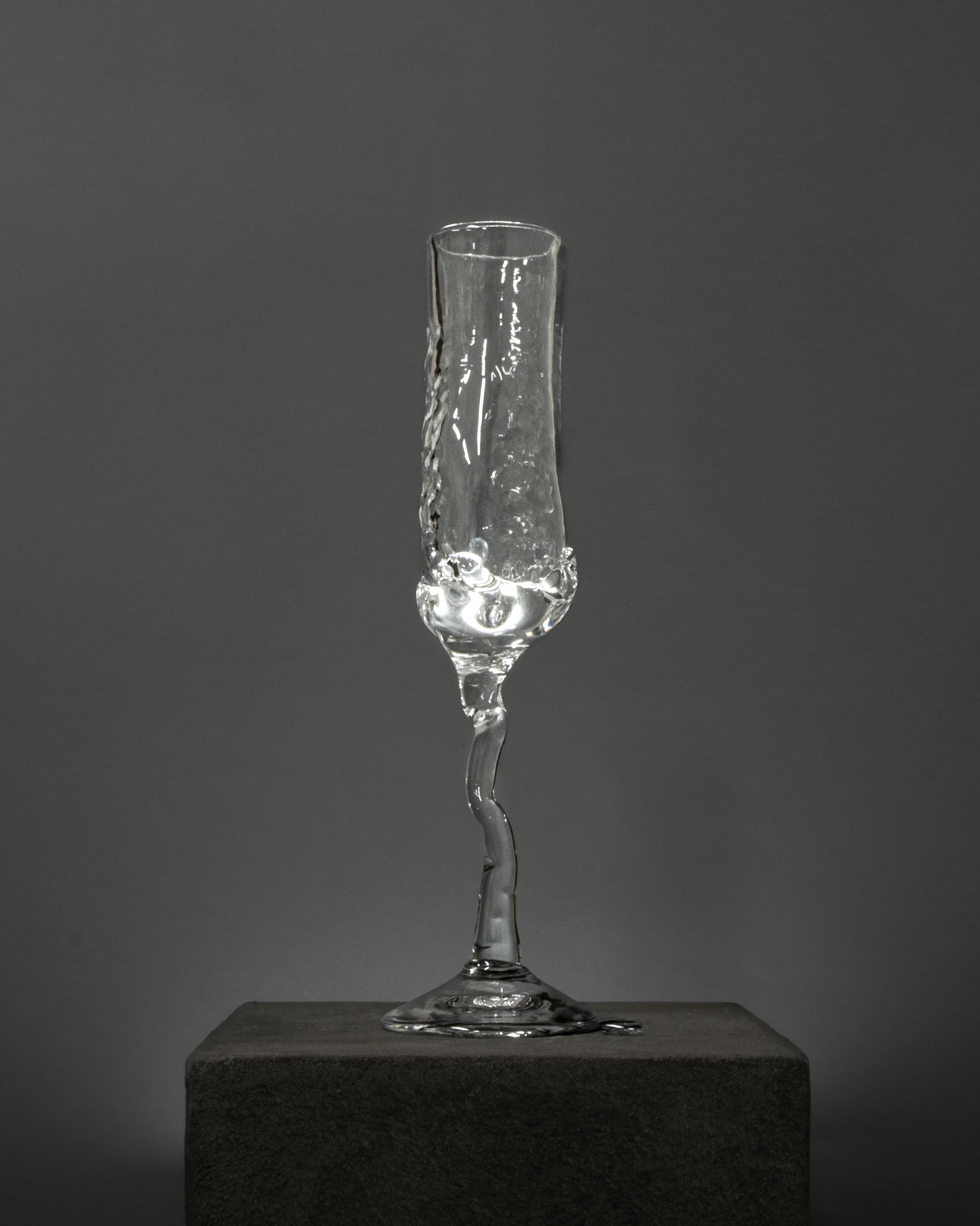 The Organic Flute Champagne Glass by Szkło Studio is a clear, hand-crafted glass with a twisted, uneven stem and an irregular, molten look. It stands on a dark block against a plain gray background with a small amount of liquid inside.
