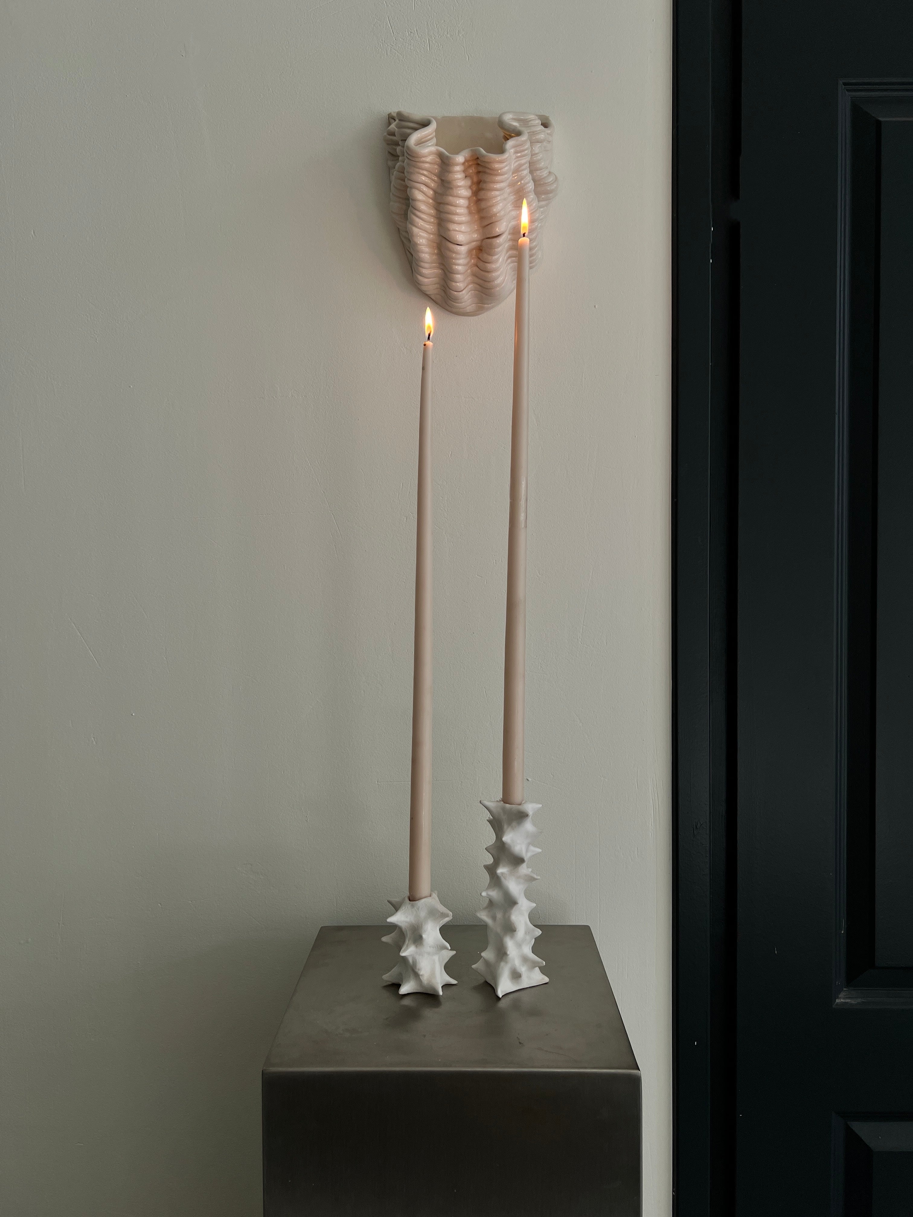 Two tall taper candles in matte white 2222Studio Spiky ceramic candle holders sit on a minimalist metal pedestal. Above, a textured organic-shaped ceramic wall piece is mounted on a light wall near a black door frame.