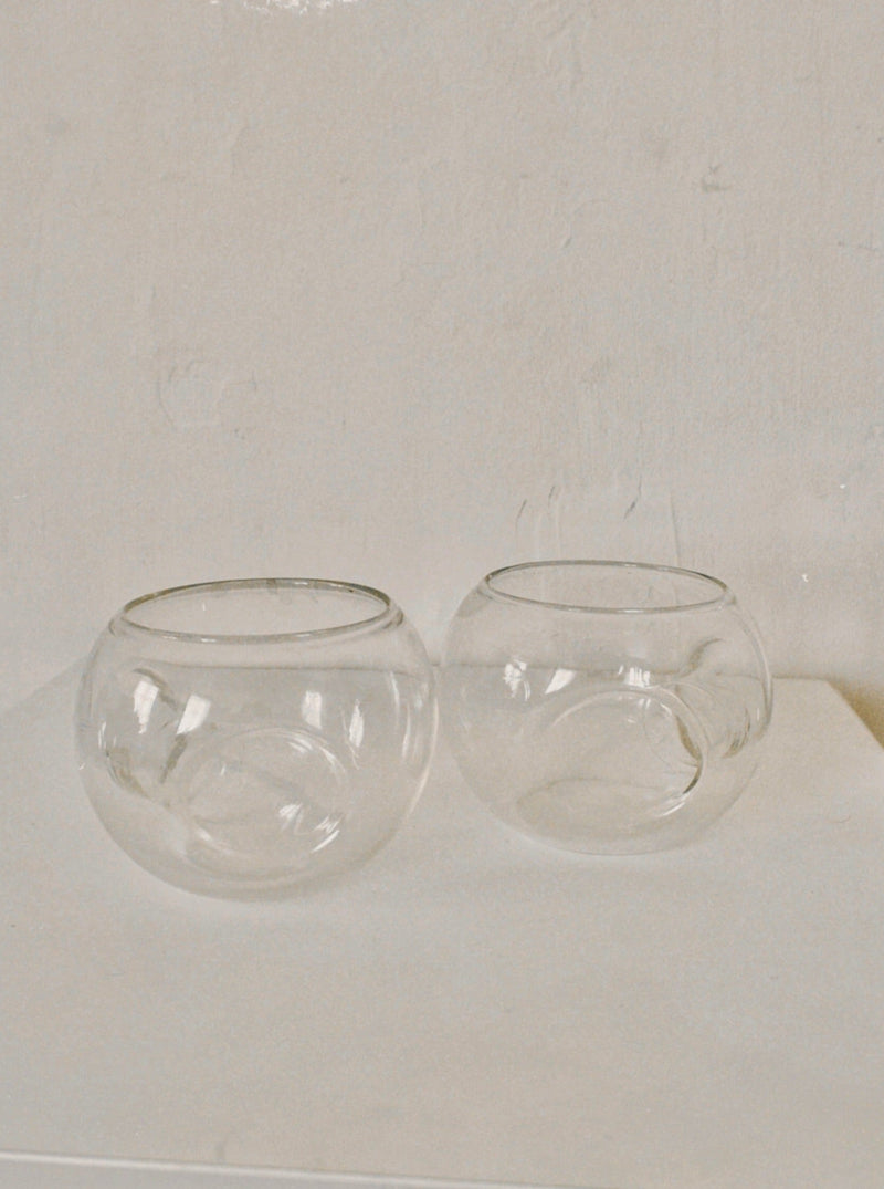 The Set of 60s Italian Glasses by Manushi features two clear, round bowls with subtle indentations, exuding elegant simplicity on a white surface against a light textured wall.