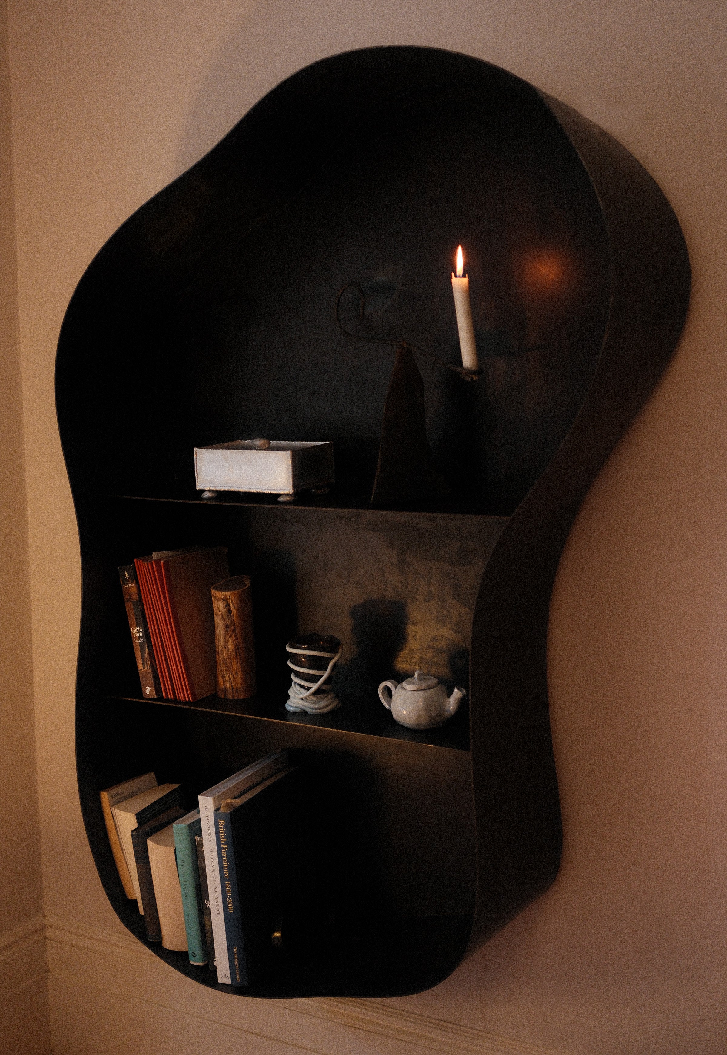 Black curved wall shelf with candle holder displaying books and teapot