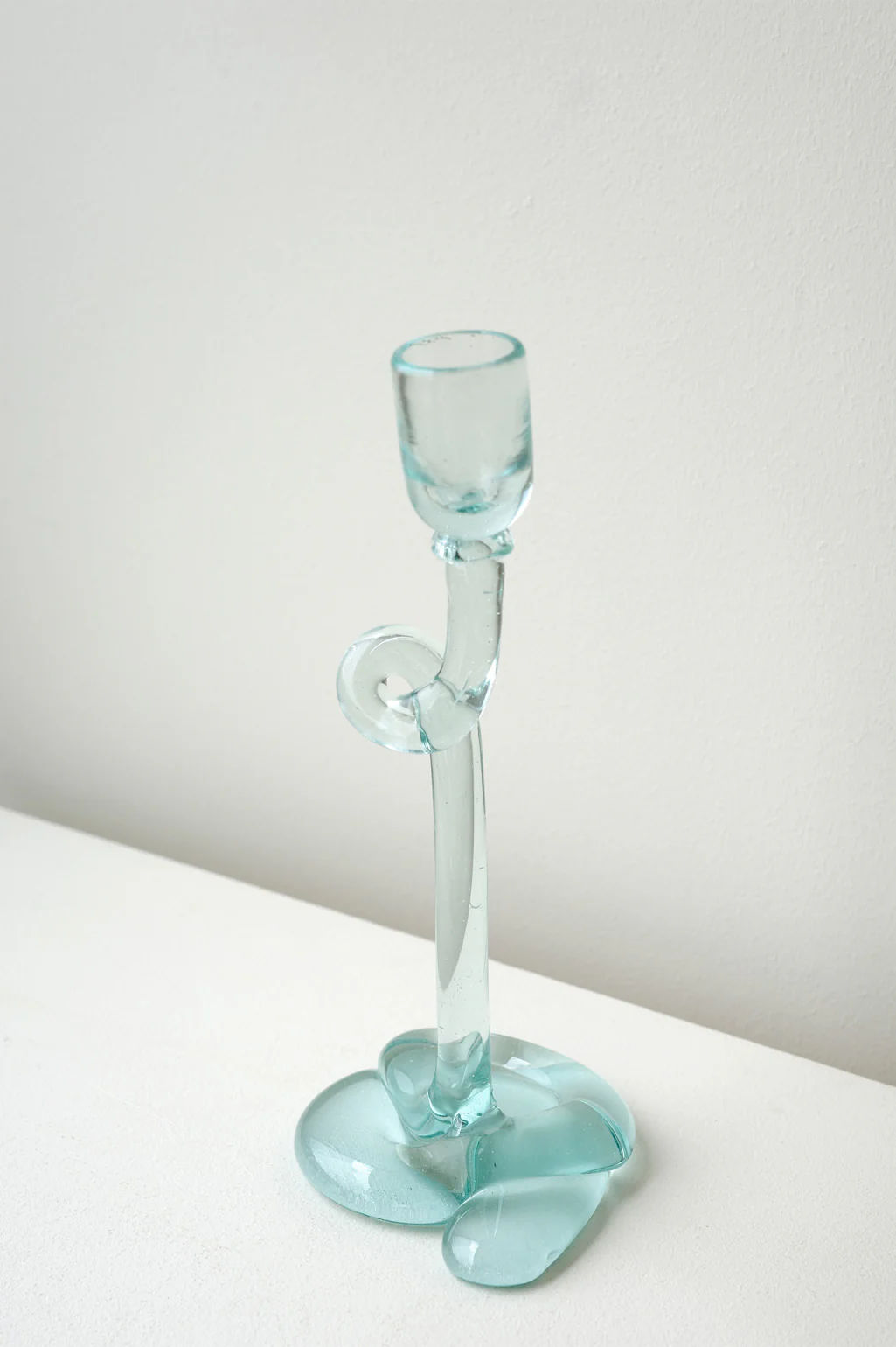 The Thaw Recycled Glass Candleholder in Clear by Completedworks features a sculptural, aqua-blue wavy design with a curved stem and fluid base, shown on a white surface against a light wall.