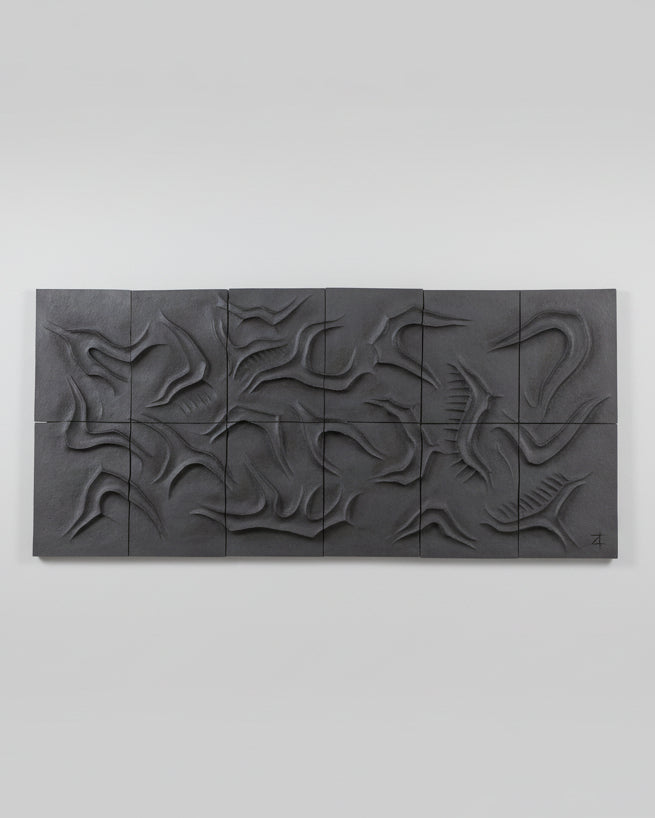 The Caldeira Wall Sculpture by Franck Scala, featuring dark ceramic and organic swirling patterns, leans against a white wall on a wooden floor. A wrought iron accent appears in the corner, enhancing this unique decorative piece.