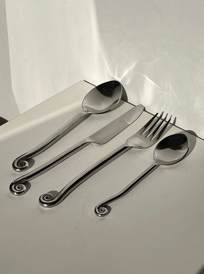 The Manushi Set of Vintage Swirl Cutlery, including two spoons, a knife, and a fork with spiral-designed handles, is arranged on a white surface. The polished metal softly reflects light.