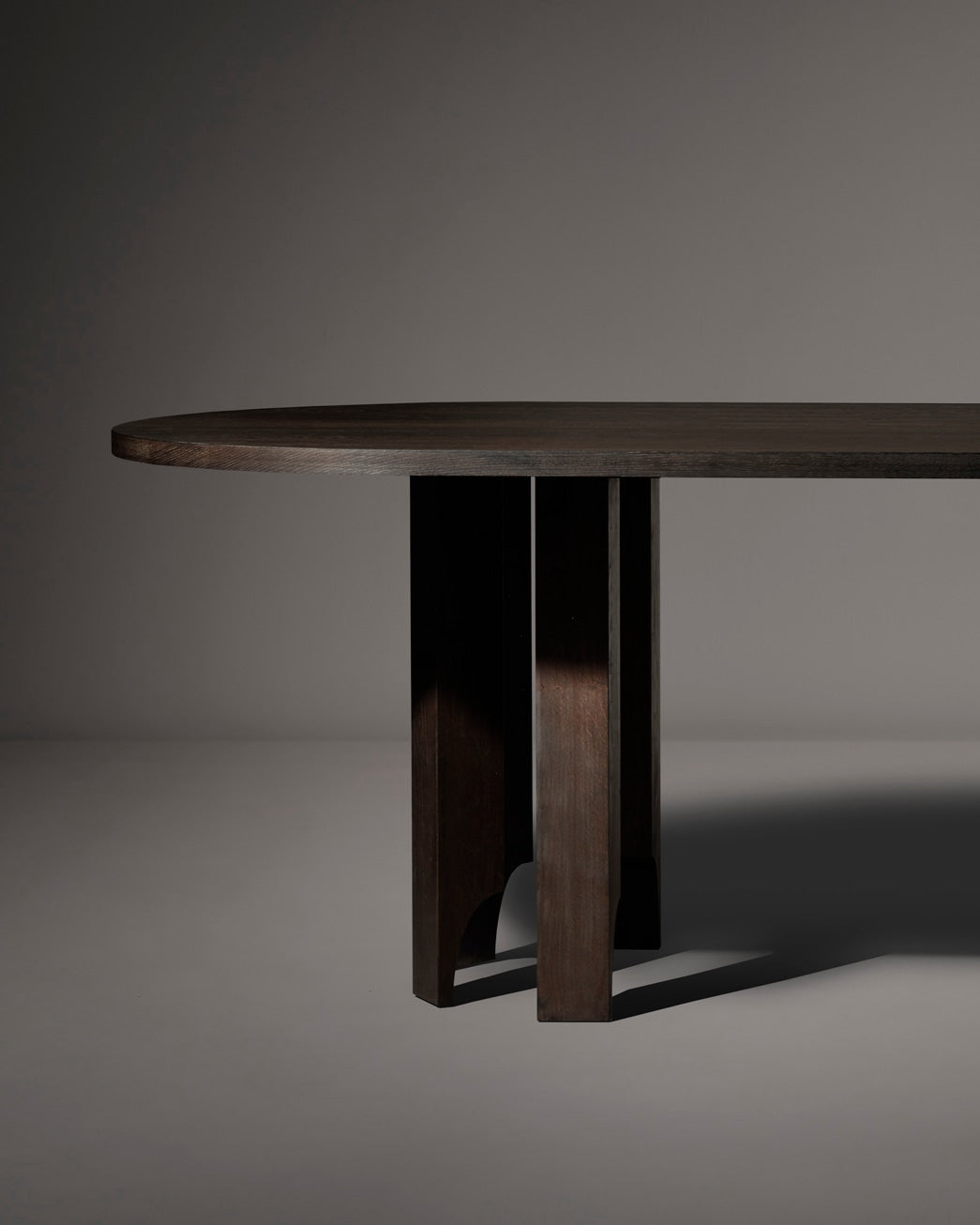 The Mokko Acer Capsule Table, crafted from solid European oak, features a thick, rounded top and sturdy intersecting legs. Its customizable design and rich texture stand out against a neutral gray background.