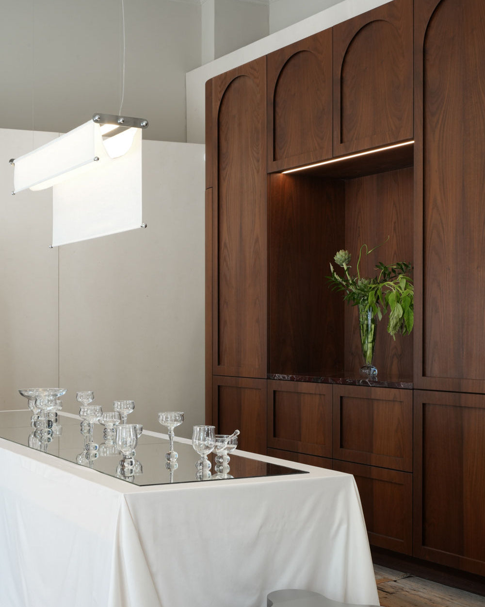 A modern dining area features a wooden built-in cabinet, a mirrored tabletop with a white cloth, glass candle holders, and a tall vase with greenery beneath the adjustable Vento Pendant by Caia Leifsdotter.