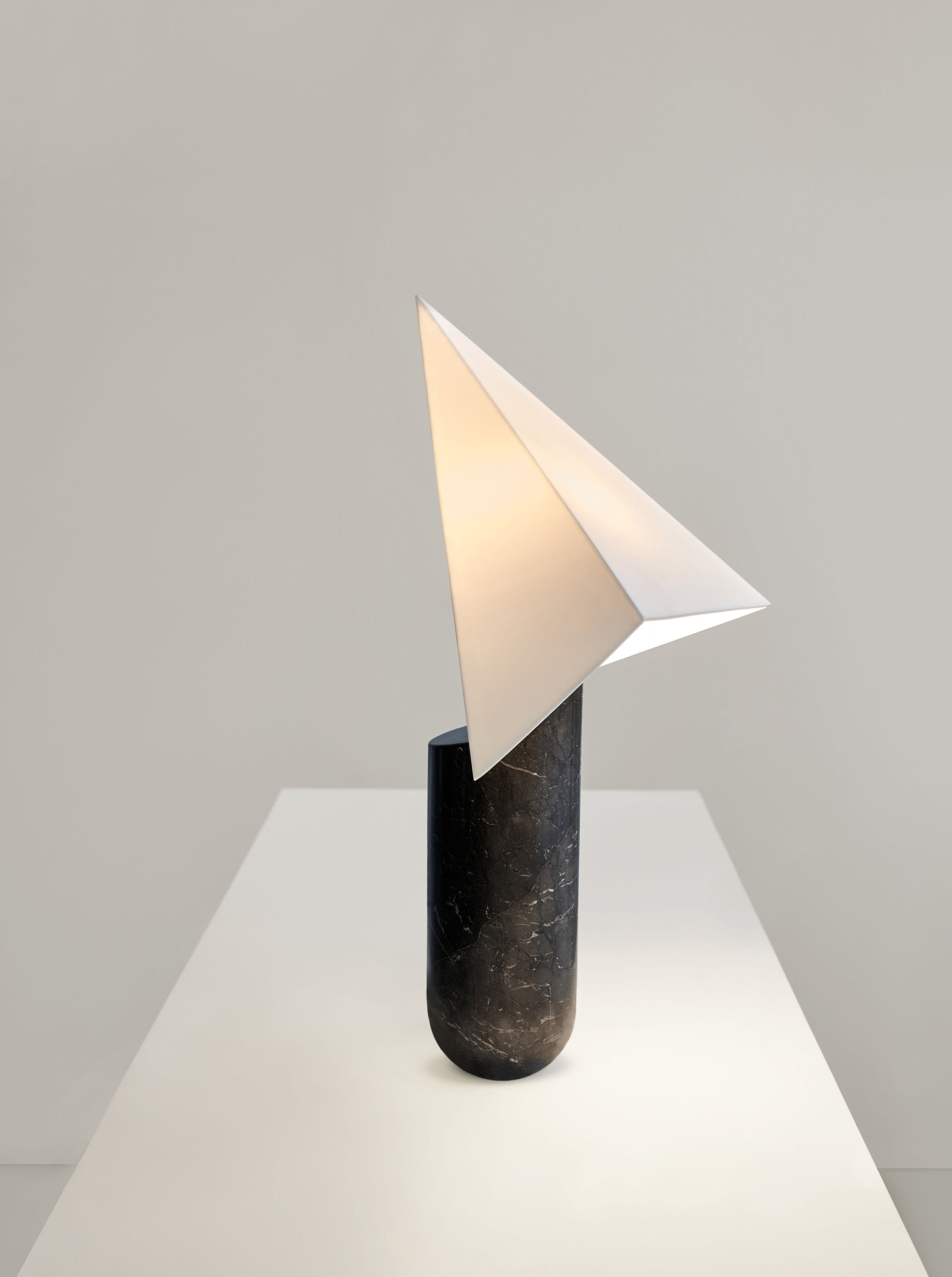 The Acme - Greek Marble Black table light by Mariza Galani features a black cylindrical Greek marble base and a white, paper plane-shaped shade, emitting a soft glow on a white pedestal against a plain background.