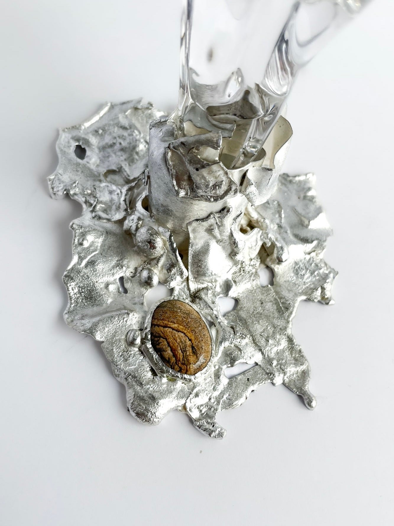 A close-up of Szkło Studio’s Jewelled Goblet no. 5 by Lindsey Fontijn shows an irregular silver base with a wood-textured stone, supporting a clear hand-blown glass piece, set against a white background.