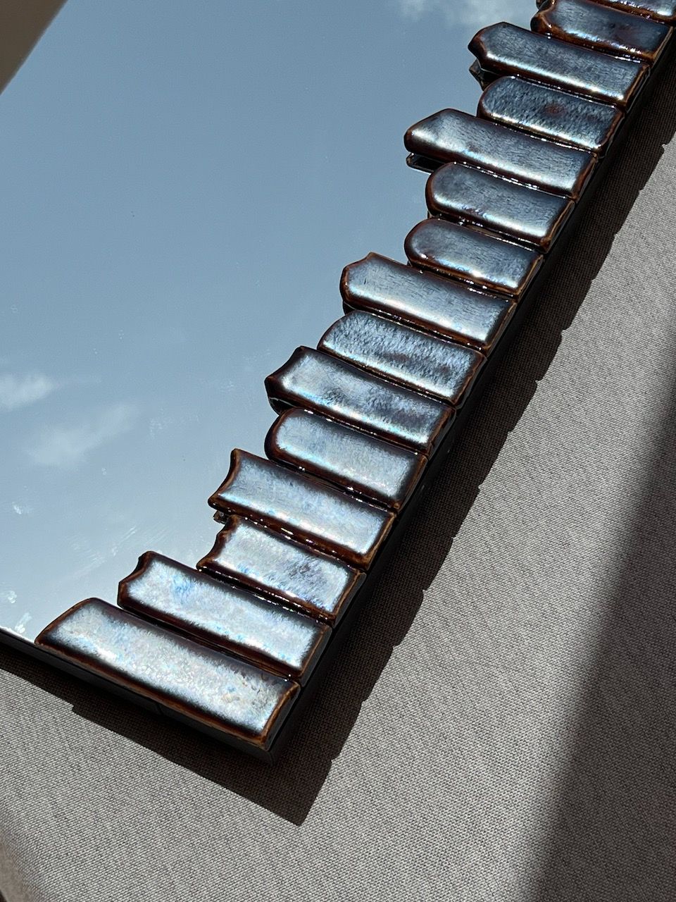The Cliff Collection from Katia Langeard features contemporary ceramic mirrors with wavy edges. Resting on a light gray textured surface, this tall mirror catches sunlight and casts soft rightward shadows.