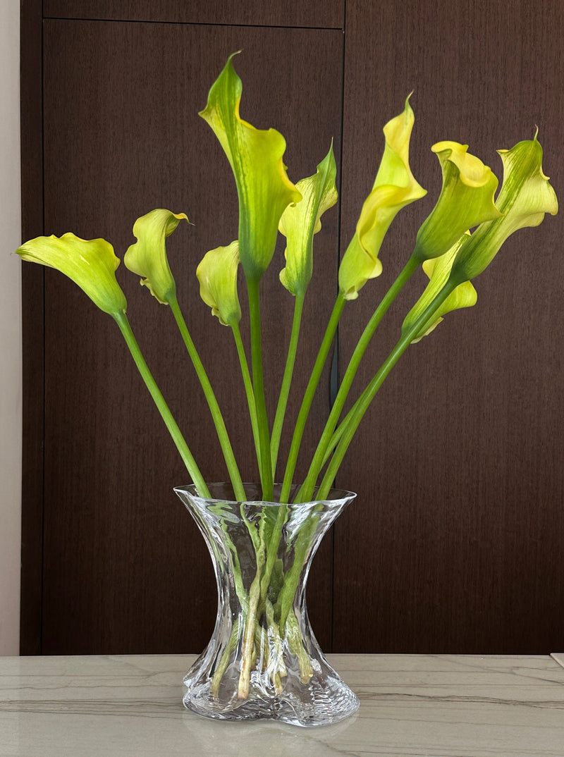 The Fvorm Tulpe Tulip Vase in Clear Glass showcases long-stemmed yellow calla lilies with green tips, arranged on a light surface against a dark wooden backdrop.