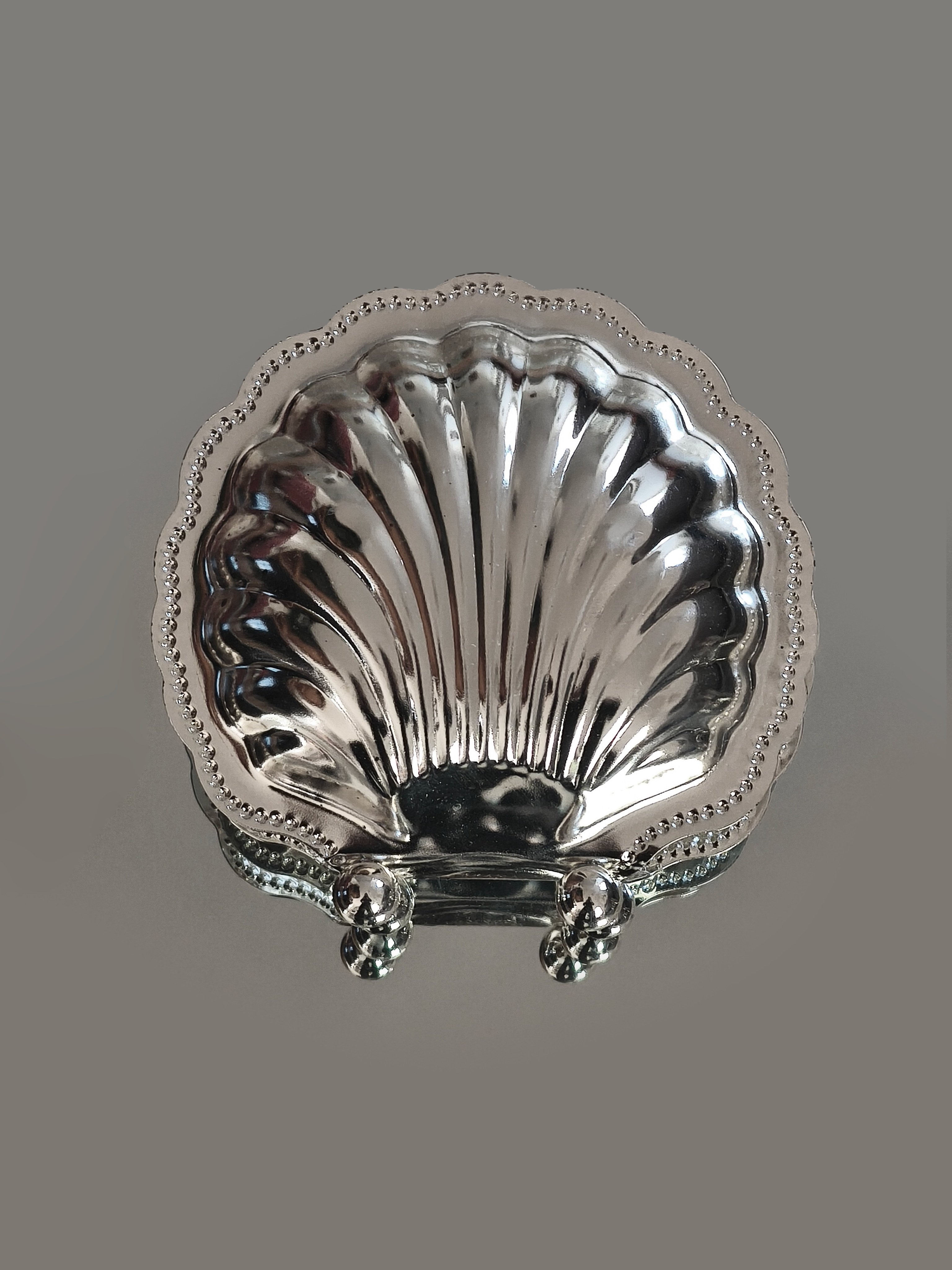 The Les Objoies Napkin Shell Holder features a silver-plated, scallop shell design with a beaded edge and three rounded feet—perfect for bringing seaside charm to your table. Displayed on a plain gray background.