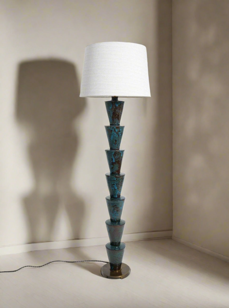 The "Nizwa" Floor Lamp by Barracuda Interiors, featuring a stacked conical teal and brown ceramic base with a white fabric shade, stands in the corner, casting a soft shadow on the beige walls and floor.