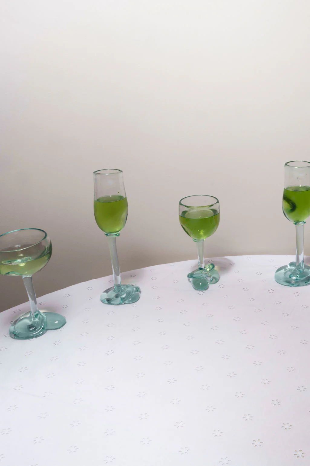 Four Completedworks Thaw - Recycled Flute Glasses in clear, each with unique shapes and heights, are arranged on a white floral-patterned table, creating a sculptural display that adds modern elegance to the setting.
