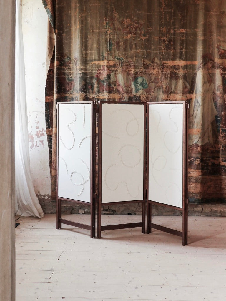 The Cavalletto Room Divider by Filippo Andrighetto, with three white panels featuring subtle abstract line art, stands on a light wooden floor before a large vintage tapestry displaying faded classical figures.
