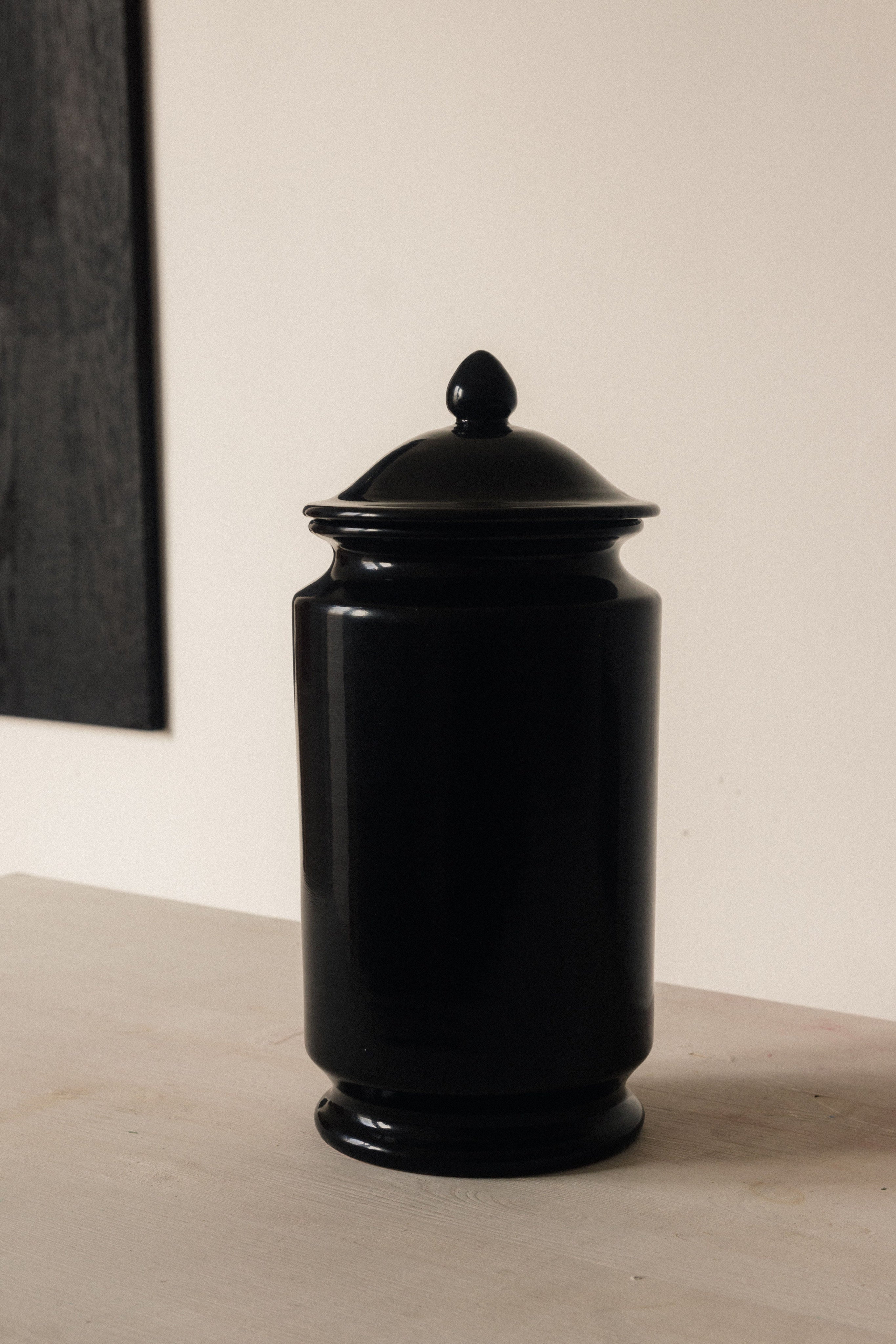 The Albarello - Minimalist Vase in Ceramic by COSEINCORSO, a glossy black handmade Italian piece inspired by traditional Albarello shapes, sits on a light surface against a beige wall with part of dark-framed art in view.