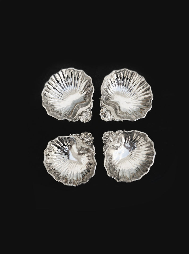 The Set of 4 small shell vide poches by Les Objoies features ornate silver-plated scallop-shaped dishes arranged in a square on a black background, each with detailed ridges and decorative accents—perfect for display.