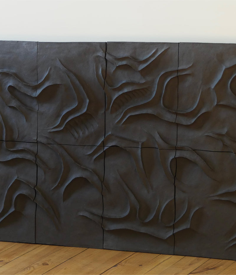 The Caldeira Wall Sculpture by Franck Scala is a dark, sculpted ceramic panel with an abstract, organic relief design of swirling raised shapes and grooves, displayed against a light wall above a wooden floor.