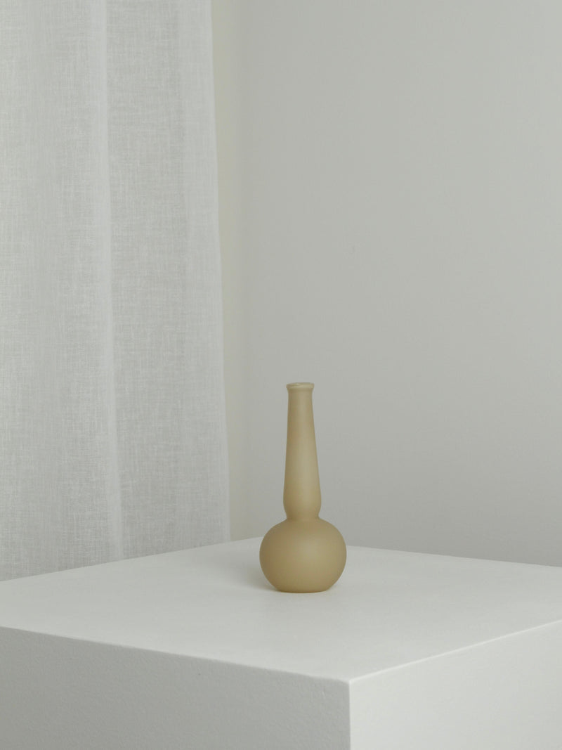 The Verre d'Onge Small Vase in Caramel, featuring a matte finish and long neck, is displayed on a white pedestal against a light wall and sheer white curtain, creating a minimalist, uncluttered setting.