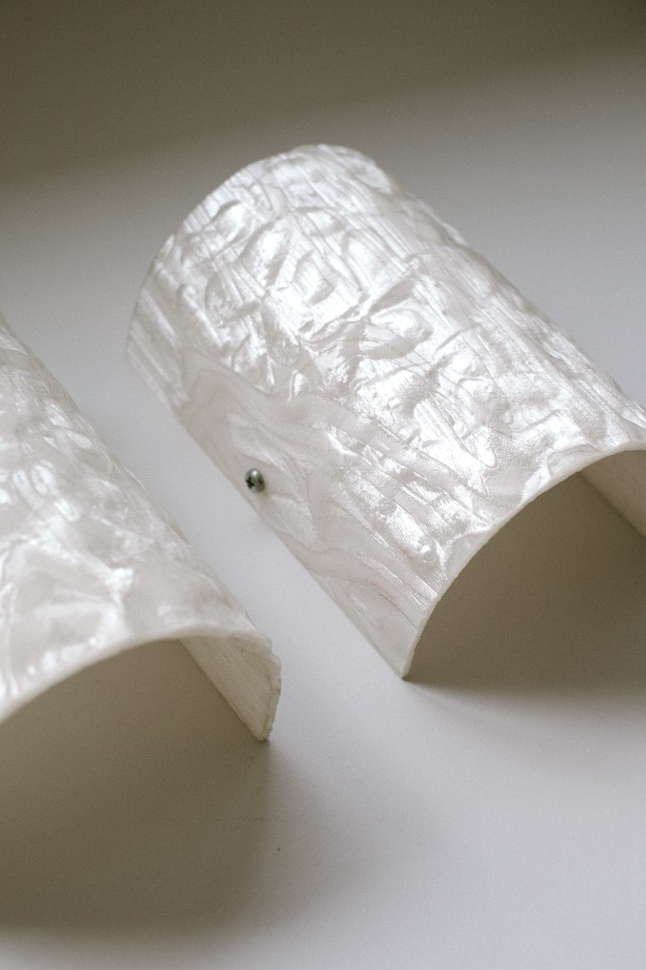 A Set of 2 Mother-of-pearl Wall Lamps by septembre studios, featuring curved white textured surfaces with a pearlescent sheen, rest on a light background. One lamp displays a small metallic screw on its side.