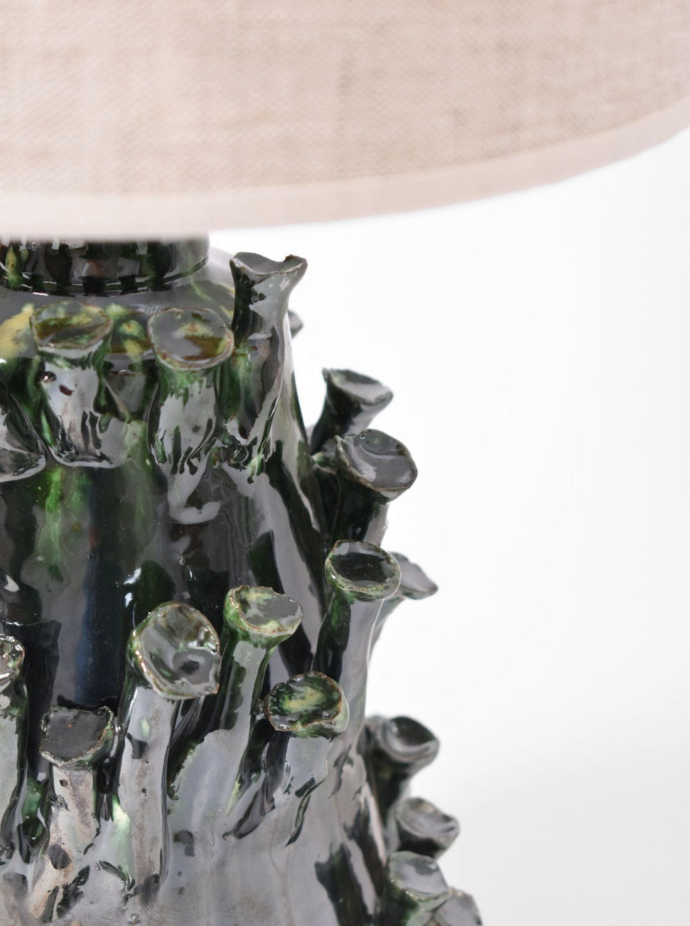 Close-up of the "Belize" Green and Black Lamp by Barracuda Interiors, a ceramic base with glossy dark green glaze and textured, organic branch-like details. Made in Portugal, it’s shown with part of a light beige lampshade at the top.
