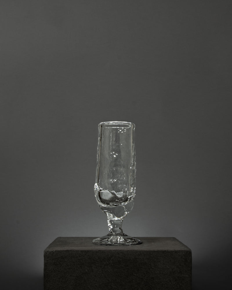 The Table Goblet by Szkło Studio, featuring a clear glass body and textured stem crafted with glass blowing techniques, is displayed on a dark rectangular pedestal against a plain gray background.