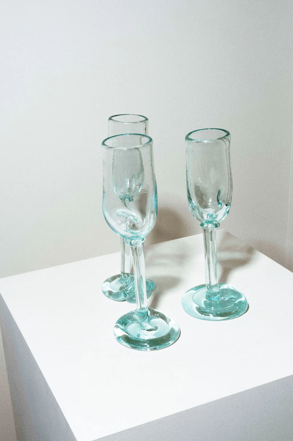 Four Thaw - Recycled Flute Glasses in Clear by Completedworks, each with a thick, uneven base and crafted from repurposed glassware, are displayed on a white pedestal against a plain white background.
