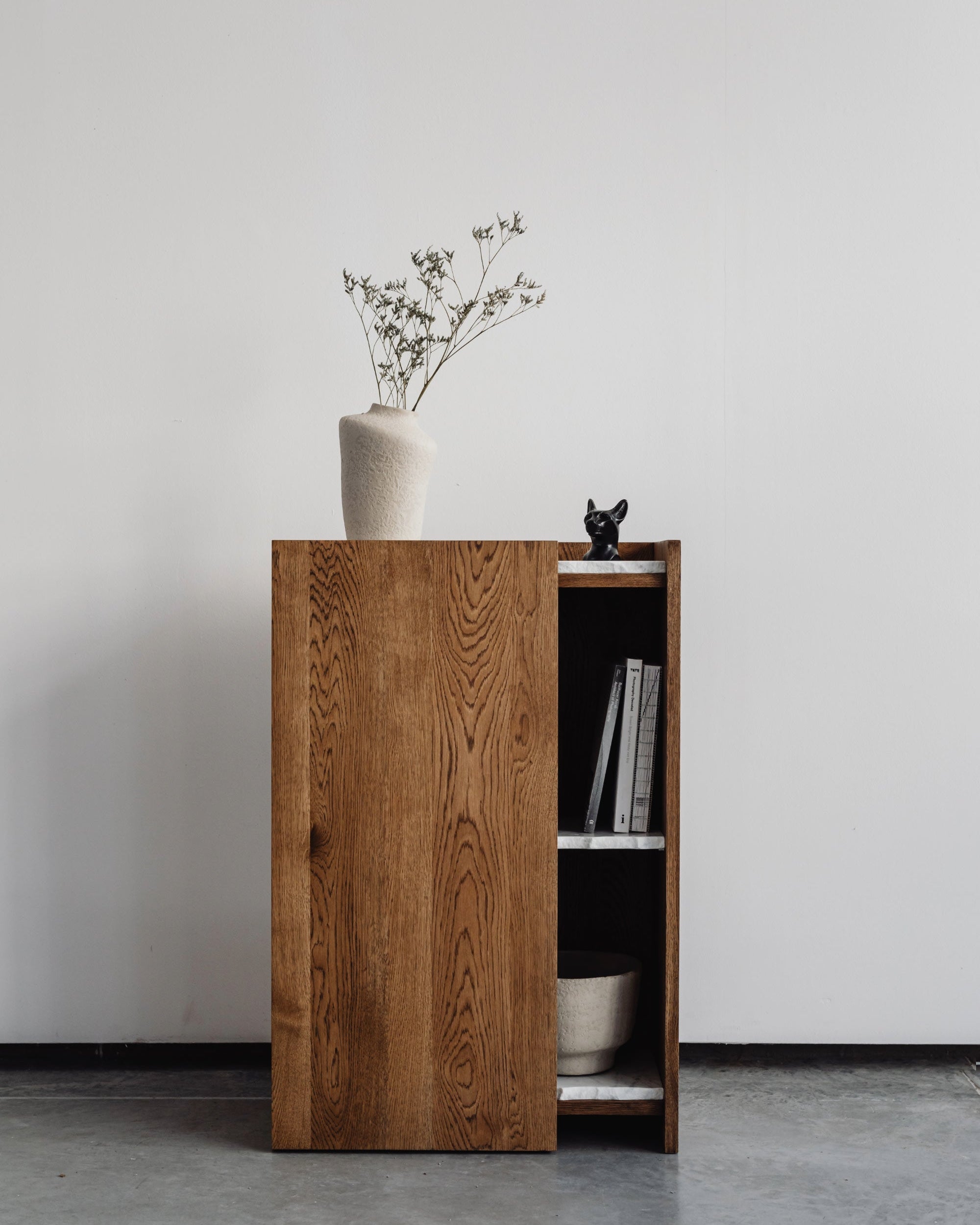 The PAVO Cabinet by un'common, a minimalist solid wood storage console with a sliding door, is topped with a white vase of delicate branches. Inside, shelves neatly hold a bowl and books, all set against a plain white wall.