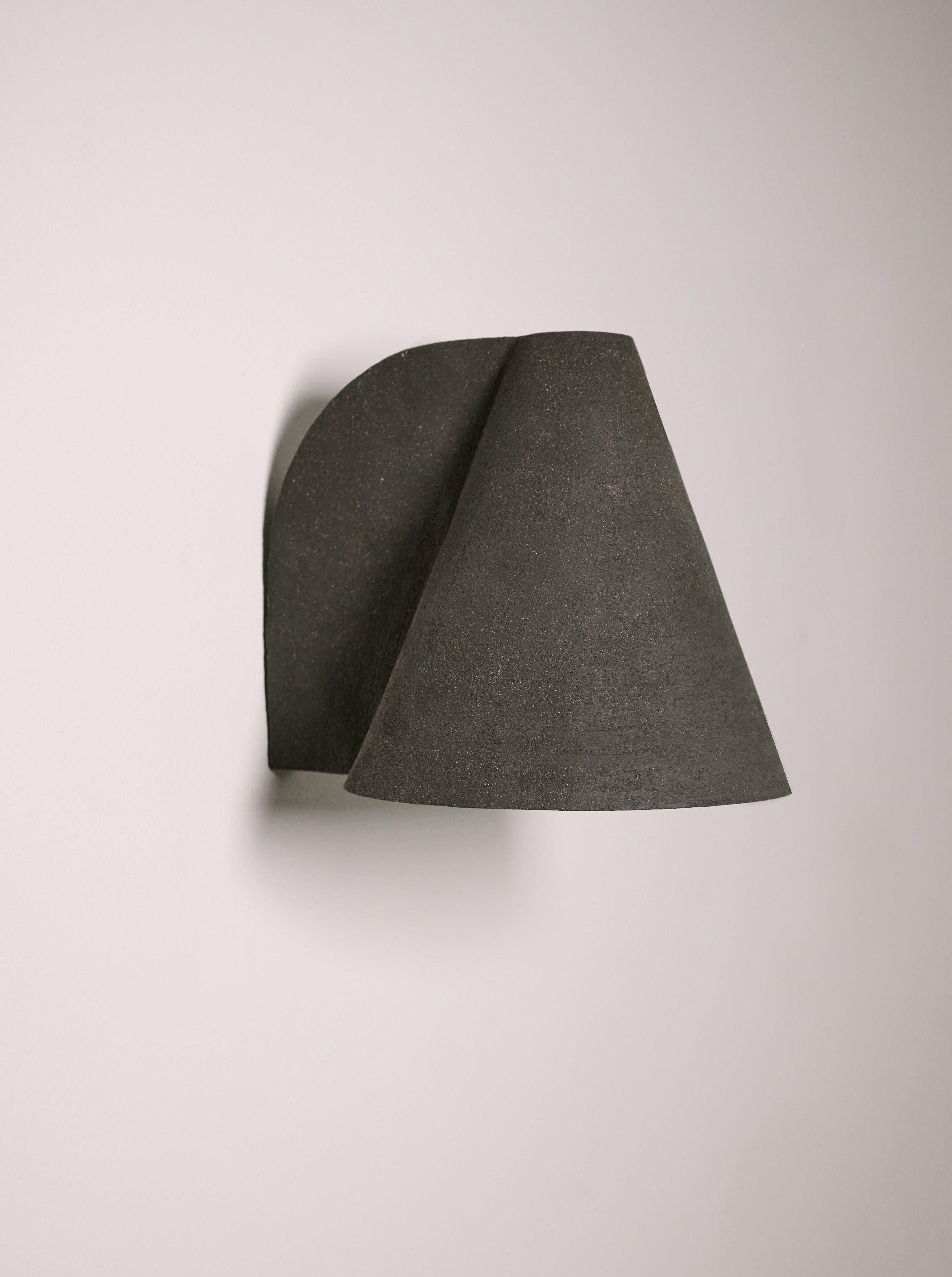The Esoteric Wall Lamp by Mariza Galani features a handcrafted stoneware, conical shade in minimalist black, mounted on a plain white wall. Its modern sculptural design combines clean lines with a unique granular texture.