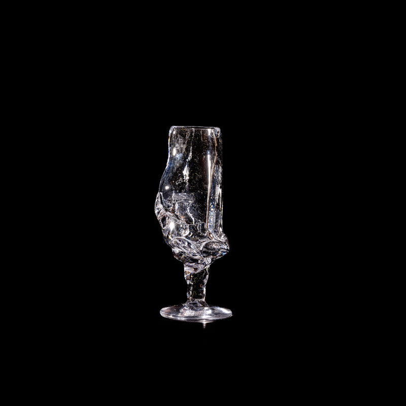 The Table Goblet by Szkło Studio, with its distorted, irregular shape and transparent glass, recalls the art of glass blowing—resembling a melted wine glass against a black background.