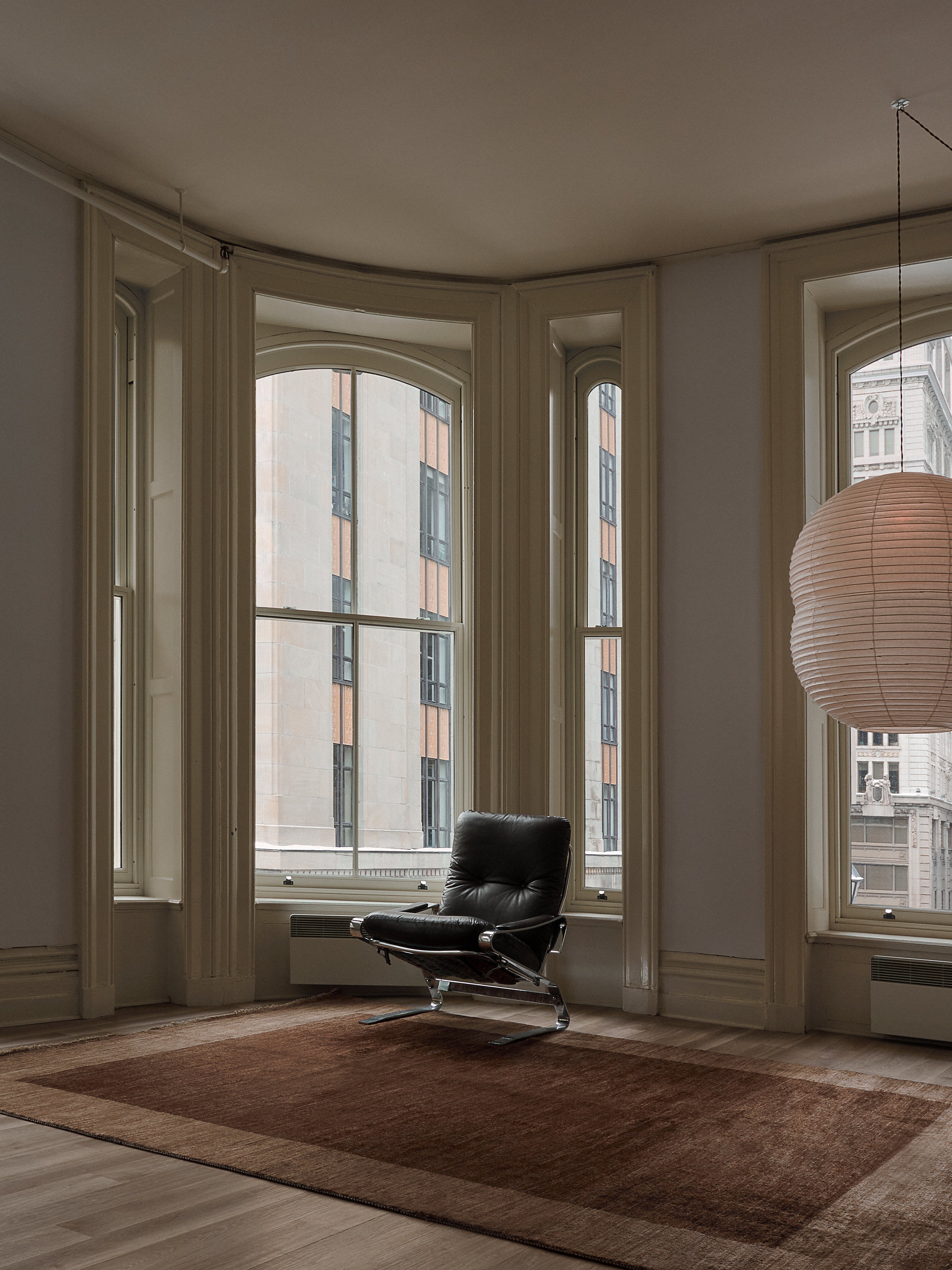 Tall arched windows showcase a city view in a modern room. A sleek black chair sits on the catalog:three Ziegler Rug in Willow (247 cm x 327 cm), under a large round white hanging light. The neutral tones create a minimalist, cozy atmosphere.