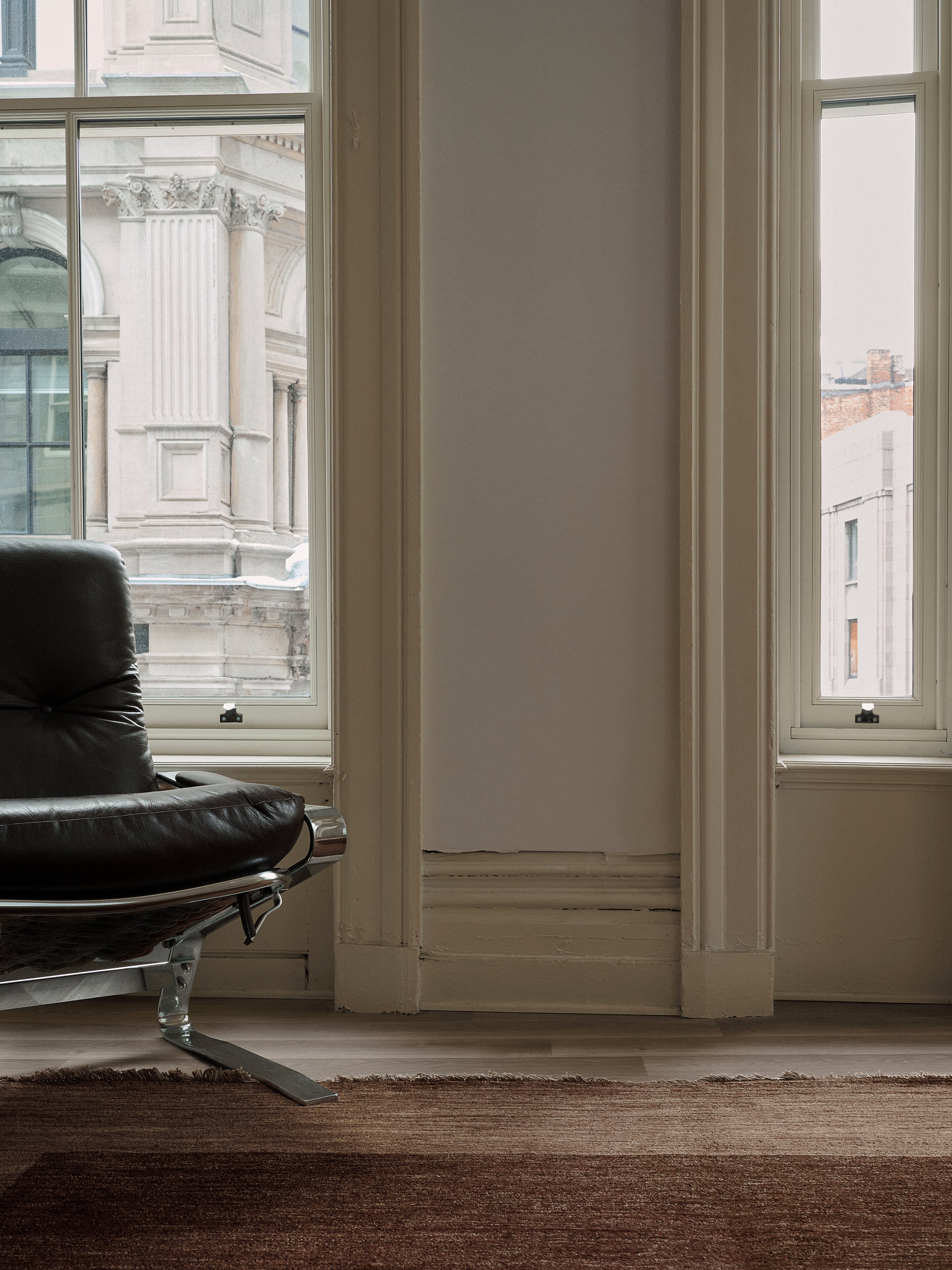 A modern interior boasts a black leather chair by tall windows with classic architecture views. The room, featuring light walls, is enhanced by catalog:three's 100% wool Ziegler Rug in Willow (247 cm x 327 cm), adding warmth and elegance.