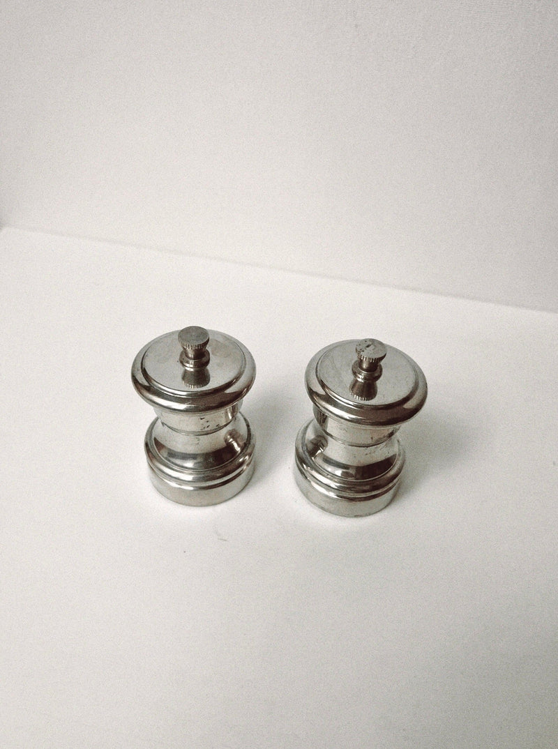 Two small, round Manushi Silver Salt & Pepper Grinders with a classic design sit side by side on a white surface against a light background.