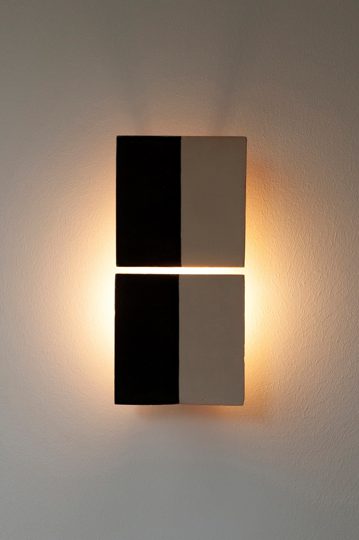 The Tiles Line N by Violaine d'Harcourt is a wall-mounted light with two square, hand-made panels divided into black and white halves, casting a warm glow and a soft halo on any light-colored wall.