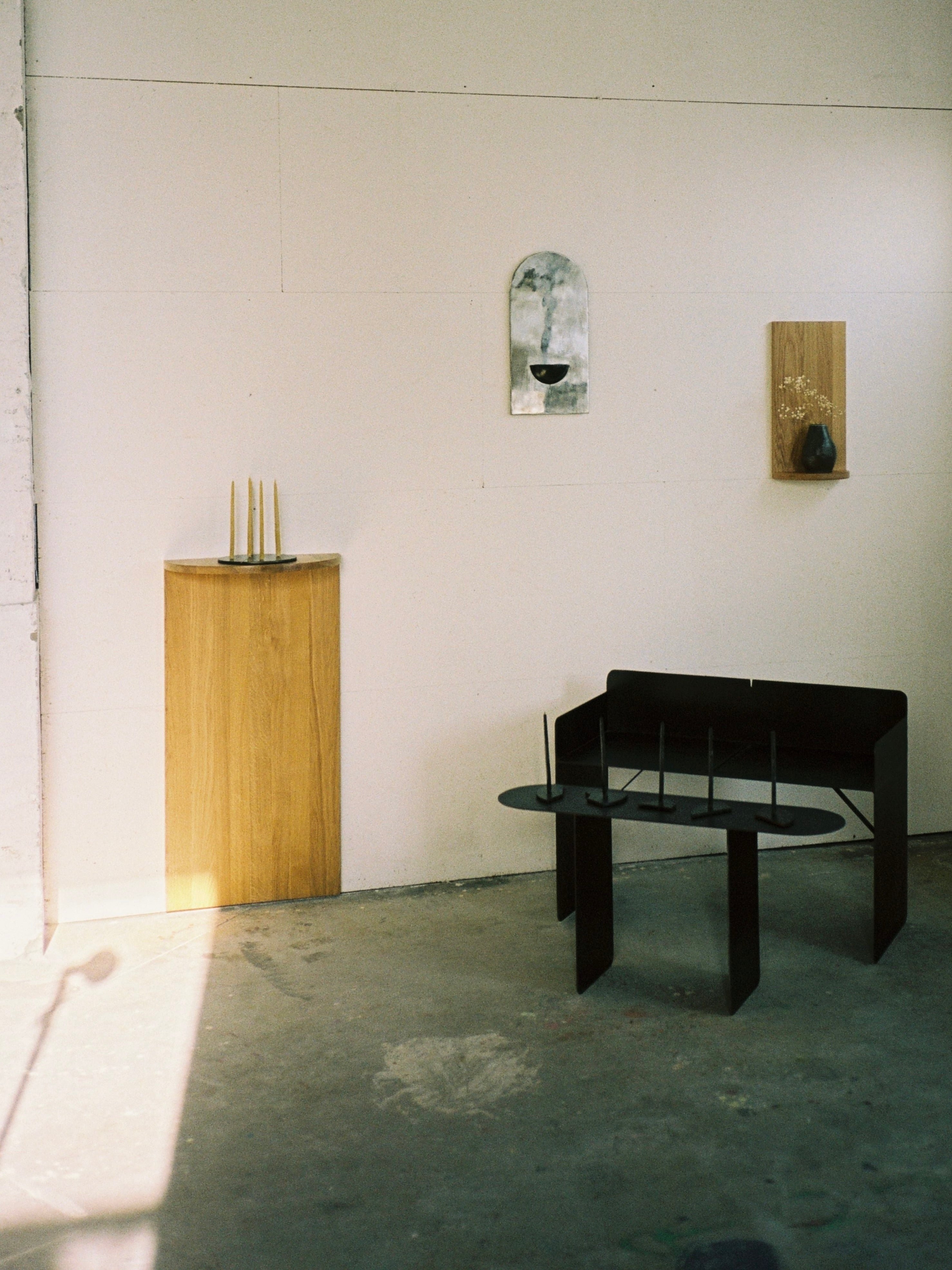 A minimalist room with white walls, a solid oak pedestal for tall candles, a modern black bench topped with black candles, COSEINCORSO's The Megaron - Minimalist Shelf in Wood (Natural Oak Large) with dried flowers, and a metallic wall mirror. Sunlight streams across the concrete floor.