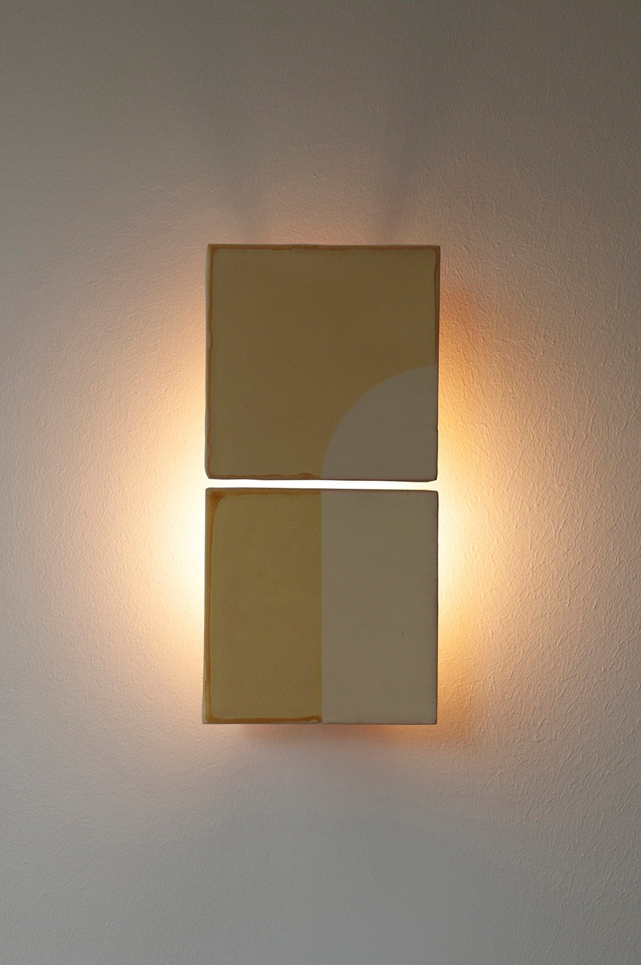 The Tiles Door J by Violaine d'Harcourt is a wall-mounted light with two stacked square panels, each displaying elegant beige and cream geometric tile patterns. Warm backlighting softly illuminates the wall.
