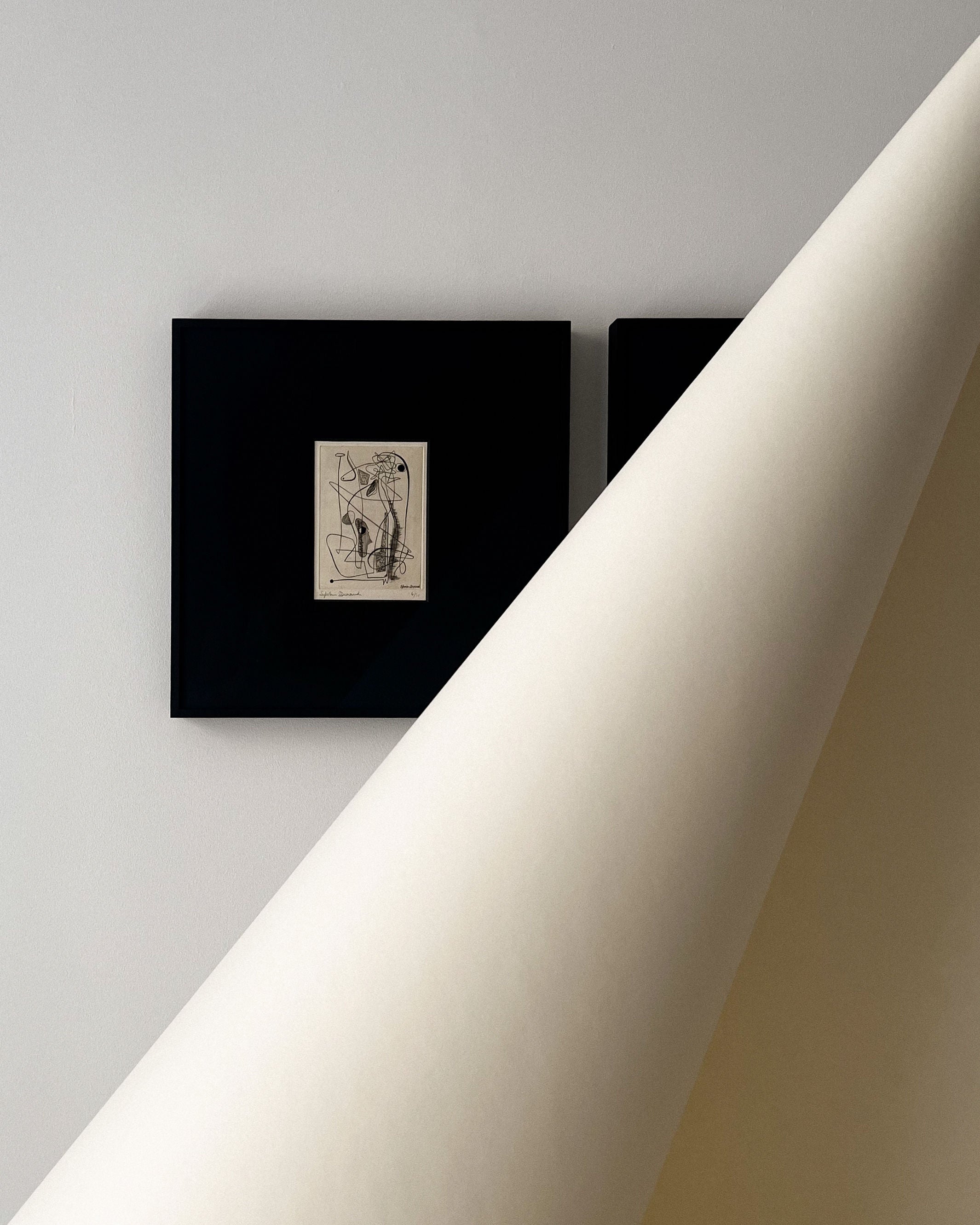 A large sheet of paper curls from the bottom right, partly covering two framed abstract black-and-white artworks on a light wall, including the "Surrealist Engraving Signed Sylvain Durand" by Collection apart.