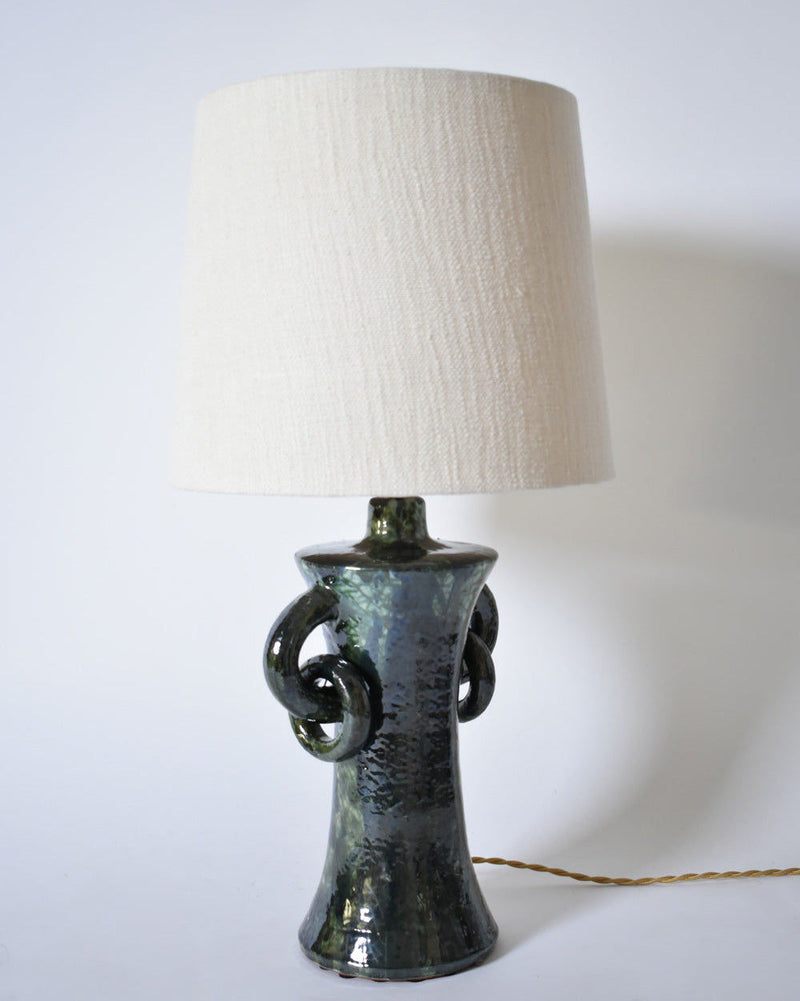 The "Kasaï" Blue & Green Ceramic Lamp by Barracuda Interiors features a textured, dark glossy base with decorative side loops, topped with a large off-white fabric shade. Made in Portugal, it is shown against a plain white background.