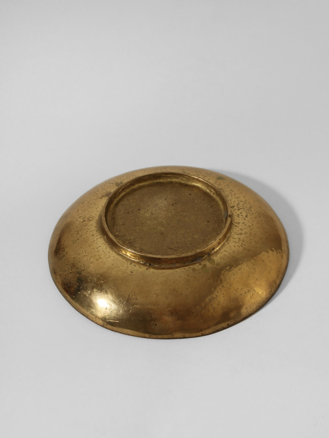 A round, shallow Massive Organic Textured Brass Bowl by Vintage Stories is shown upside down on a plain, light gray background. The bowl has a textured, reflective surface with small spots and a slightly raised rim at the base.