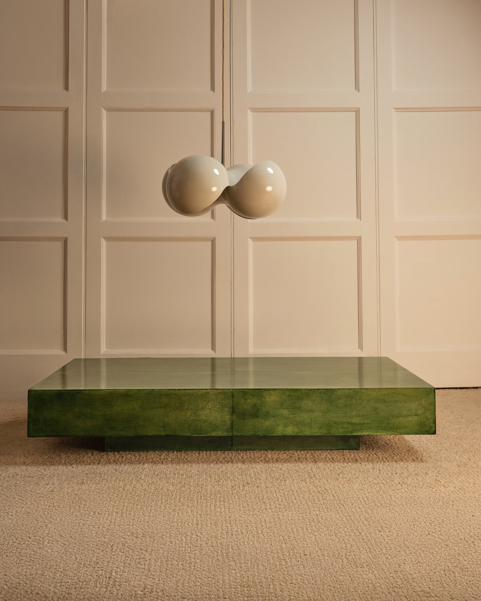 A Galerie Vauvart Green parchment table sits on a beige carpet in front of cream paneled walls, with a unique white double-globe pendant light hanging above it.