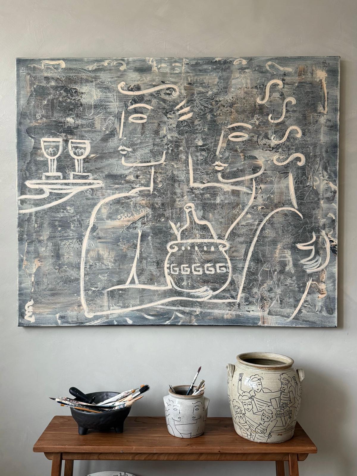 Toast to Half Full," a minimalist painting by Claire de Wolf depicting two abstract figures with drinks, hangs above a wooden table holding art supplies, a decorated mug, and a ceramic vase adorned with line drawings—true contemporary expression.