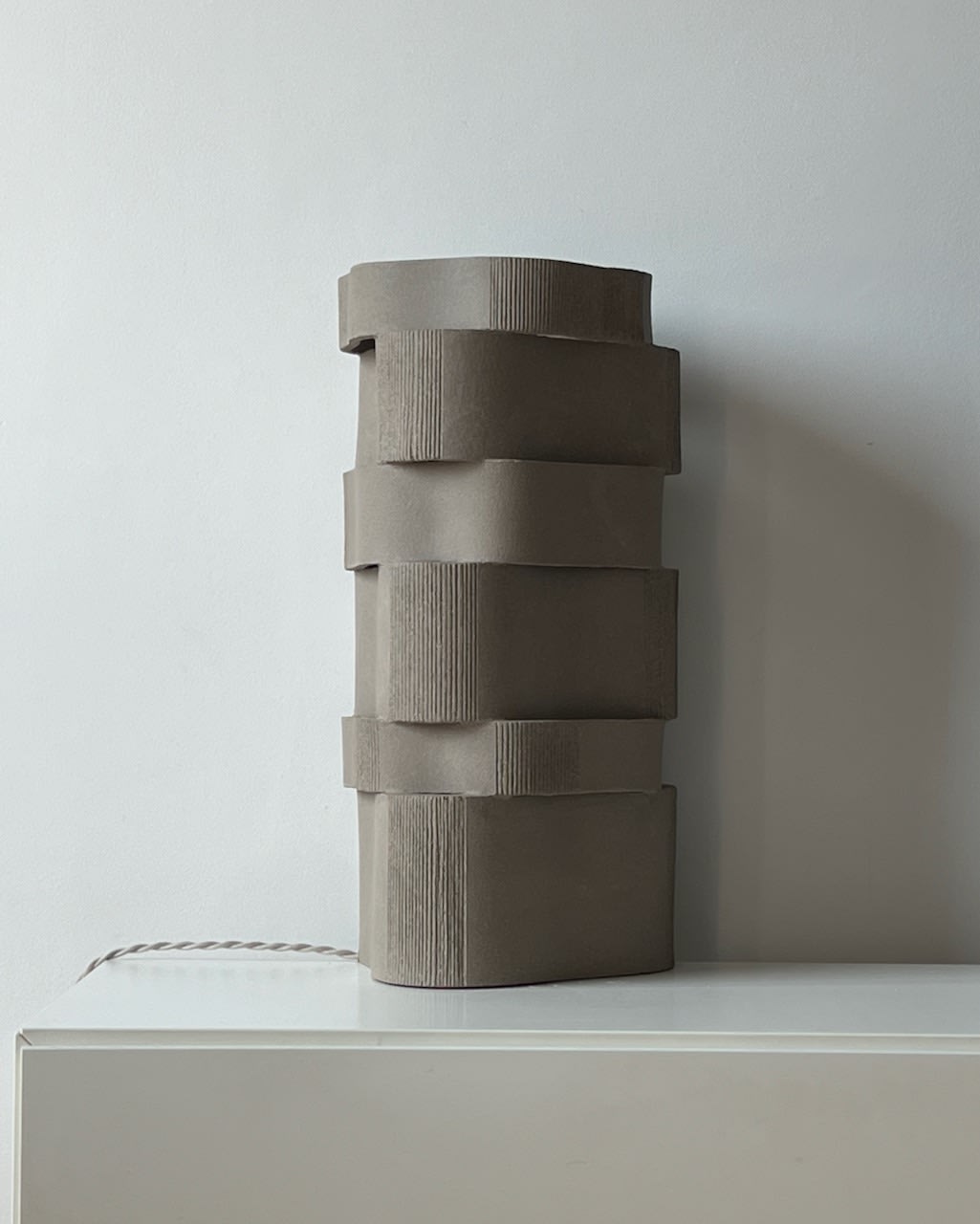 The 'Too 60' Ceramic Table Lamp by Katia Langeard, featuring a tall, modern stacked wavy matte gray design, sits on a white surface against a plain light-colored wall.