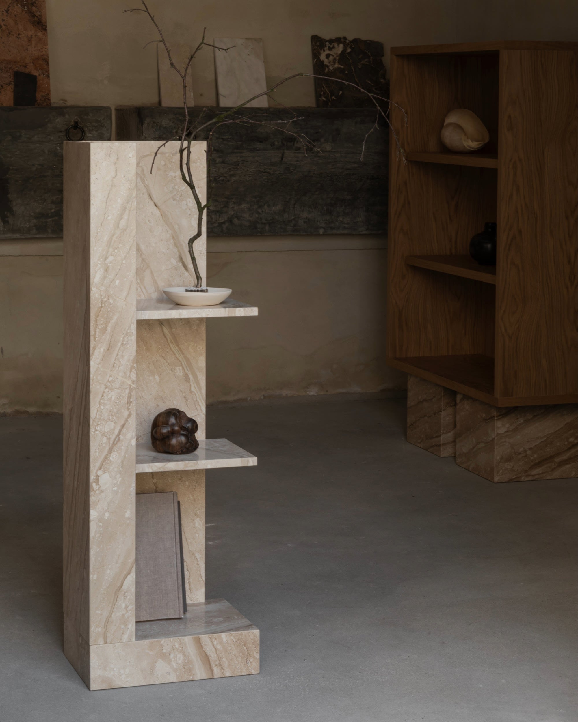 A minimal CONE Marble Shelf by un'common displays a bowl with a thin branch, a small dark sculpture, and a closed book. In the background, a wooden bookcase and textured wall panels enhance the room’s space-saving neutral décor.