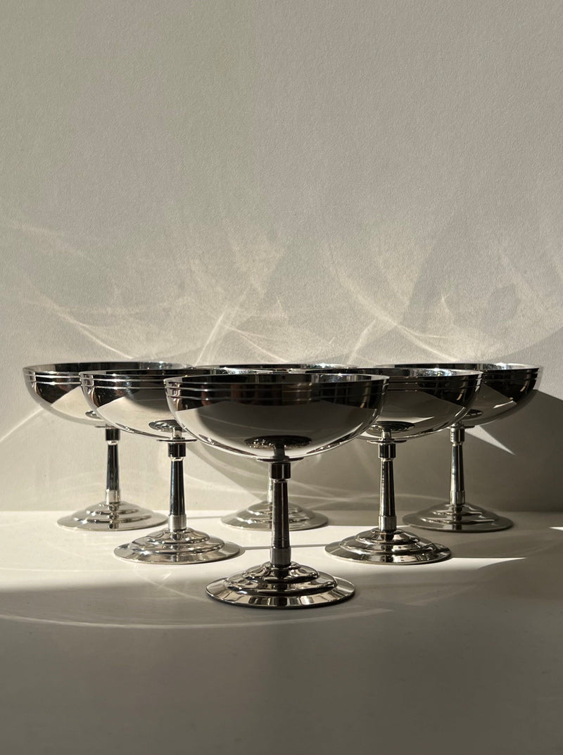 The Manushi Set of 6 Ice Cream Coupes features shiny silver dishes arranged in a semicircle on a white surface, casting soft reflections and shadows against a light-colored wall.