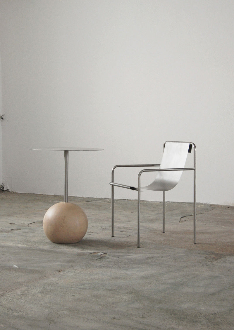 A minimalist scene showcases the SALAK DESSERT chair with a white sling seat next to a round table on a wooden sphere base, all resting on bare concrete and set against a plain white wall.