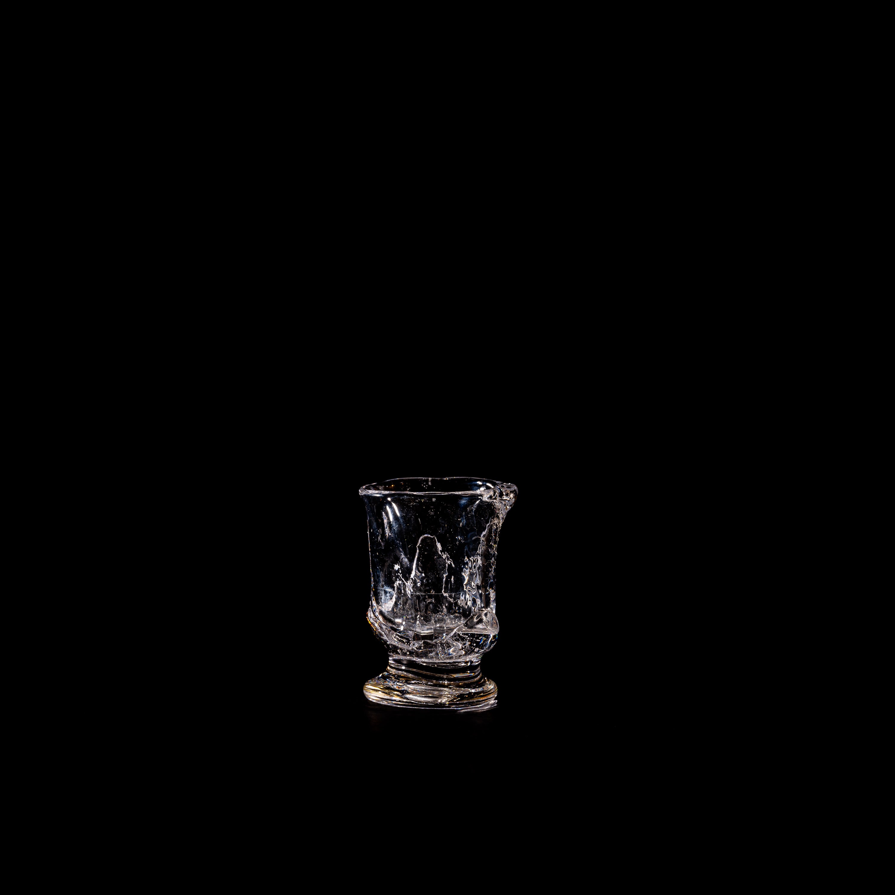 The Organic Shaped Table Glass by Szkło Studio, featuring dramatic cracks and jagged edges, stands alone on a black background under a single light—showcasing the brand’s evocative approach to glassware design.